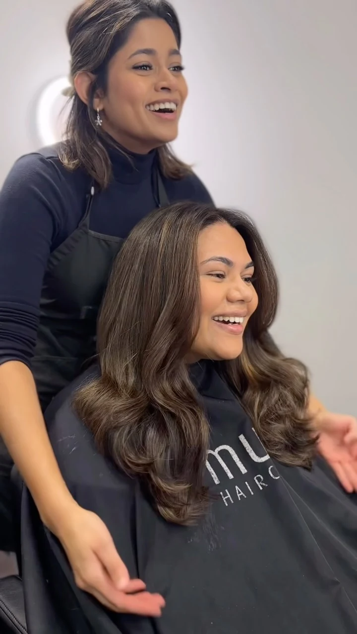 Oshi Dias in a black turtleneck and apron, laughing behind a client who loves her new glossy brunette blow-dry